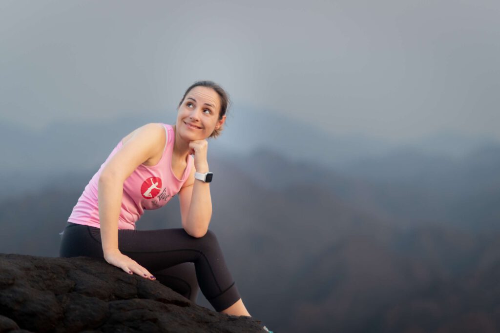 FibromFrau sitzt in sportlicher Kleidung auf einem Berg mit nebliger Landschaft im Hintergrund.