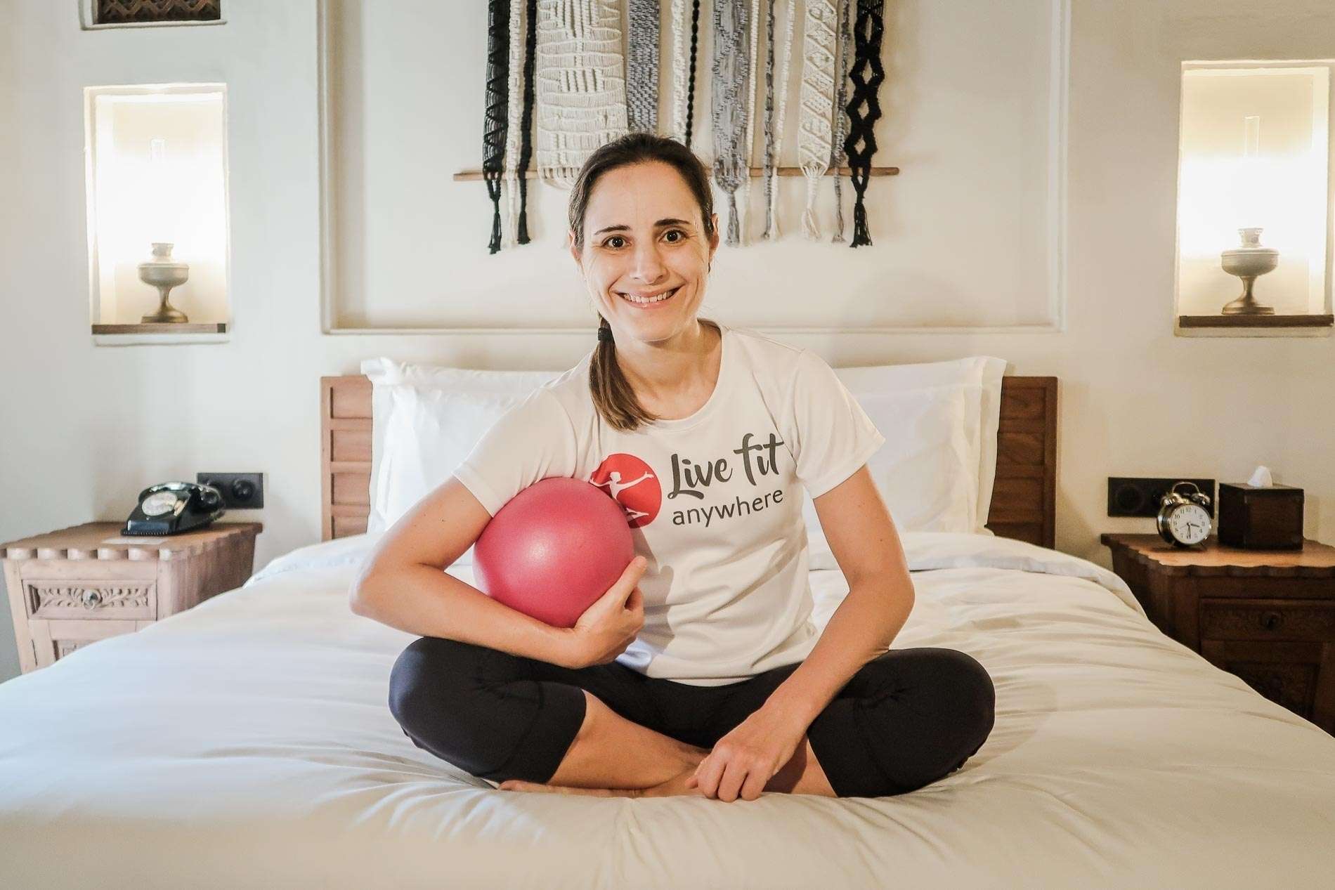 Eine Frau sitzt laechelnd auf einem Bett und haelt einen kleinen roten Gymnastikball, was auf Coaching und Gesundheit hindeutet.