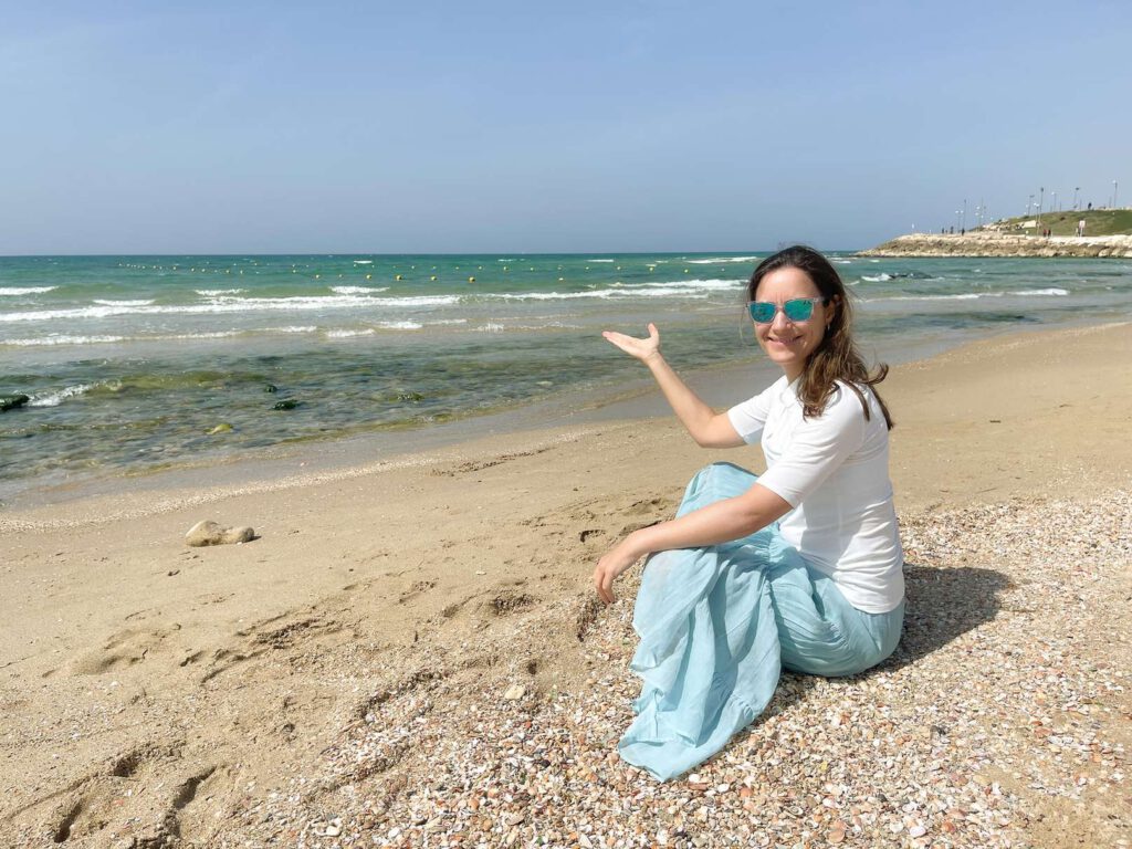 Eine Frau sitzt am Strand und zeigt aufs Meer im Kontext von Coaching und Entspannung.