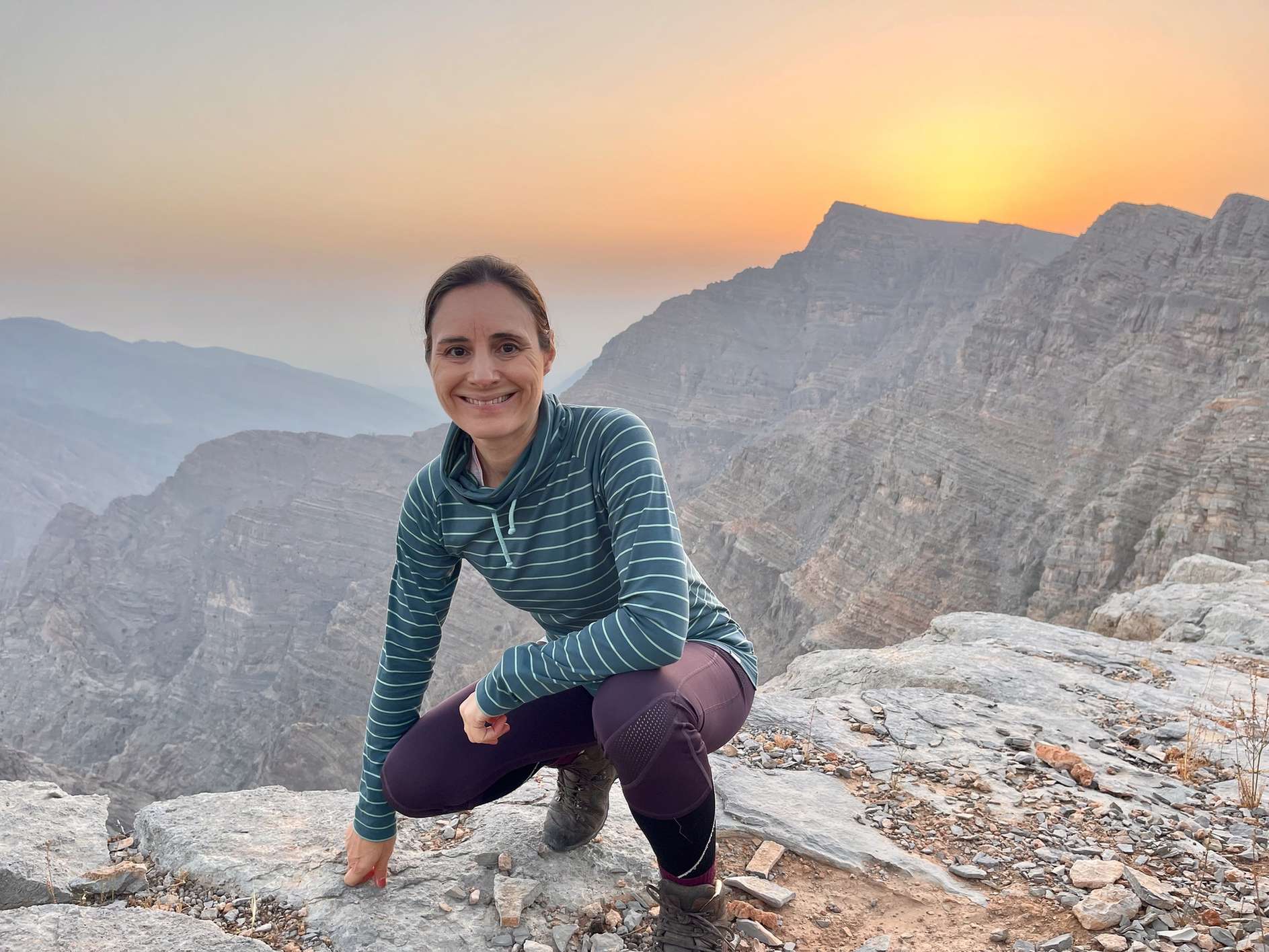 Eine Frau sitzt laechelnd auf einem Felsen bei Sonnenuntergang in einer bergigen Landschaft.