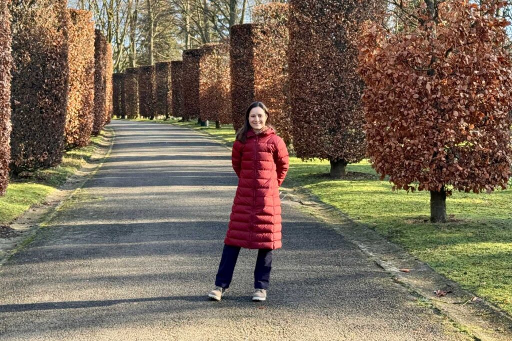 Woman in a winter coat stands in front of large trees in the park